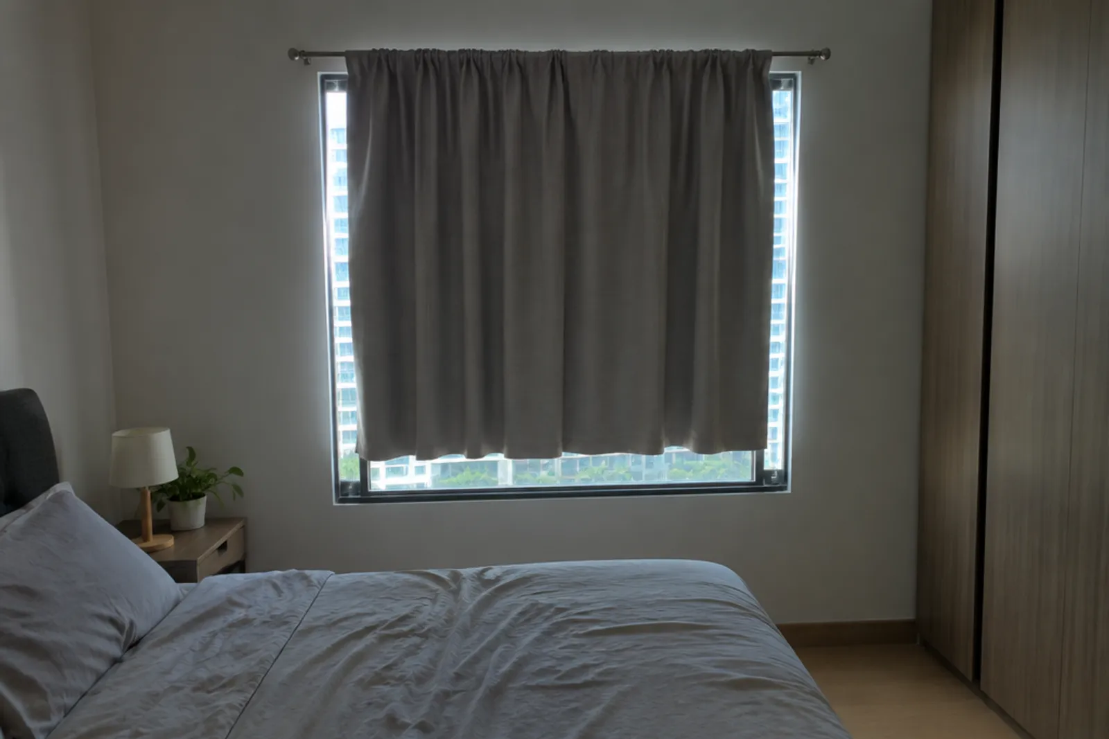 A short grey curtain on a narrow rod that leaves light leaking around a bedroom window.